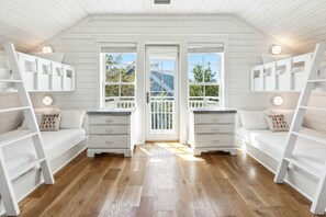 Une pièce avec un plancher en bois, des murs blancs et un balcon doté d’une balustrade. On y trouve deux lits blancs avec des oreillers et une commode blanche avec des tiroirs.