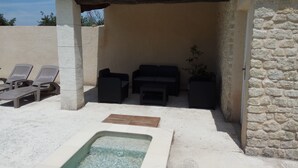 Un espace extérieur aménagé en terrasse, avec un mur en pierre, un coin salon couvert avec une table et des chaises, et un jacuzzi.