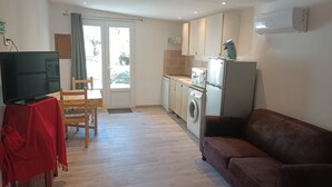 Un espace de vie compact comprenant un canapé, un téléviseur posé sur un meuble, une table à manger, un coin cuisine avec un lave-linge, et une porte-fenêtre coulissante donnant sur un espace extérieur.