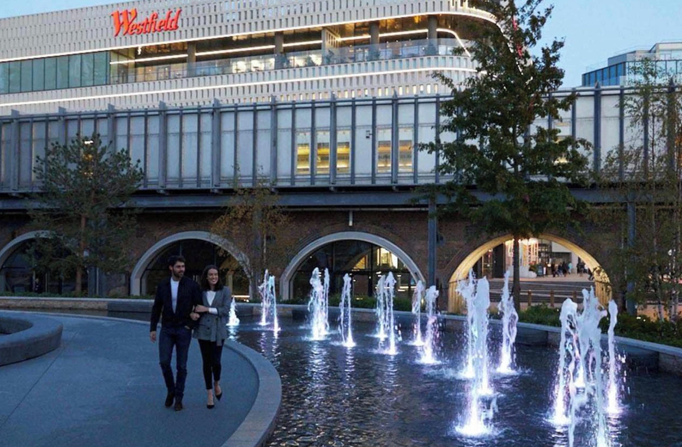 A modern building with a prominent sign, a fountain area, and a couple walking by.