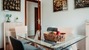 Un espace repas moderne avec une table en verre, deux chaises, un panier en osier rempli de fruits et des œuvres d’art encadrées accrochées aux murs.