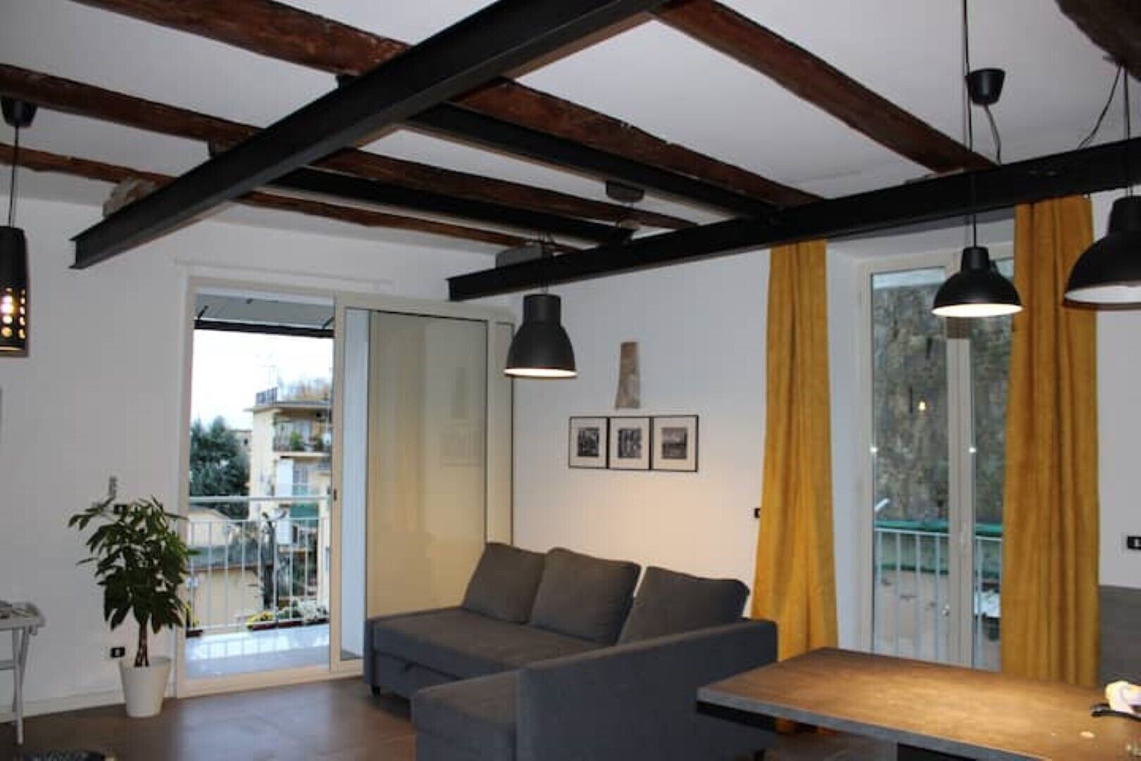 Ein modernes Wohnzimmer mit Sofa, Esstisch und Topfpflanze. Ein Balkon bietet Blick auf Gebäude, und eine Tür führt nach draußen.