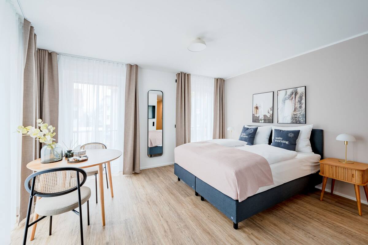 Una habitación de hotel moderna con cama, mesita de noche, lámpara y un rincón de comedor con mesa y sillas.