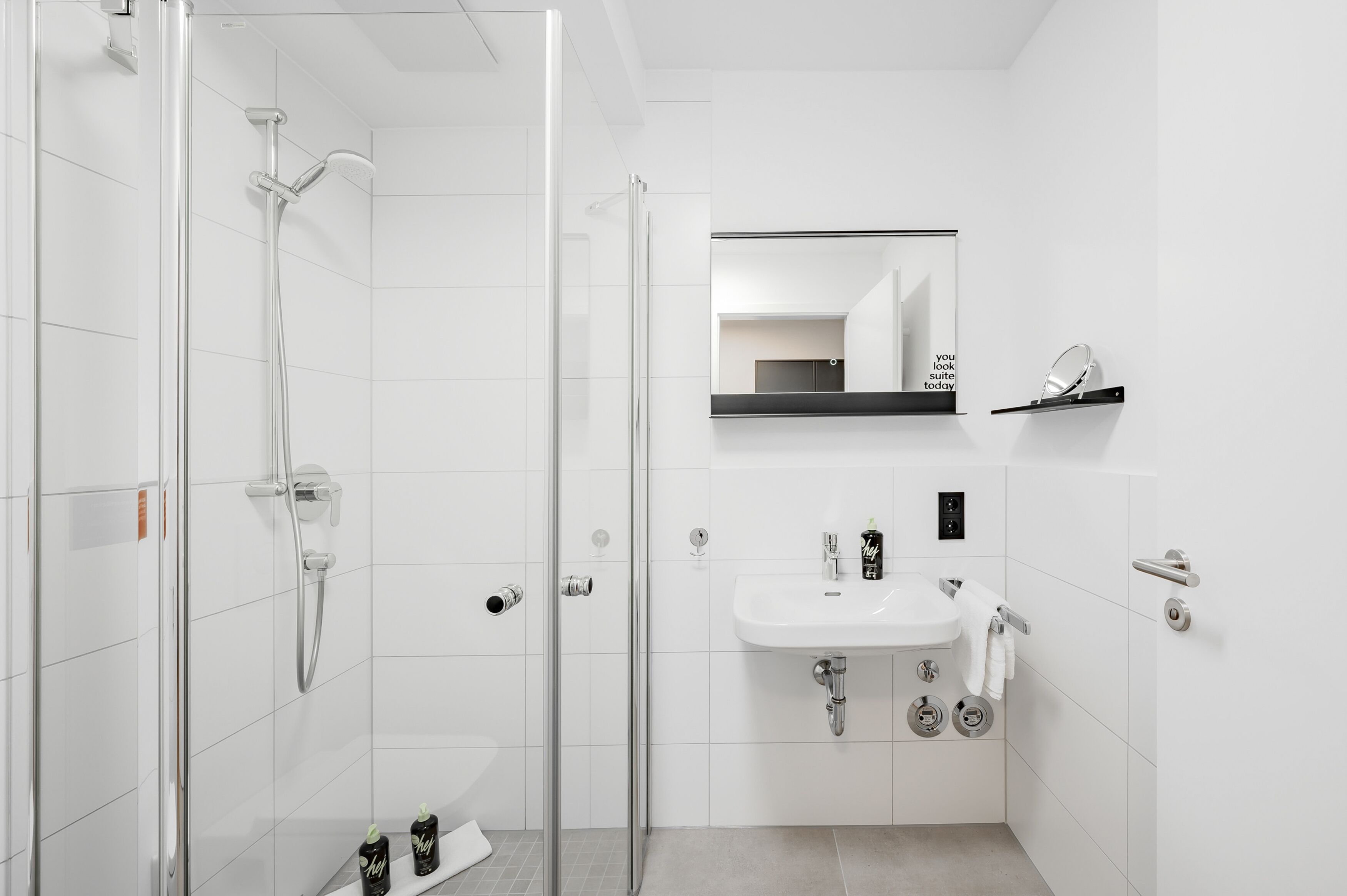 A modern bathroom with a glass-enclosed shower, a white sink with a mirror above, and a shelf with toiletries.