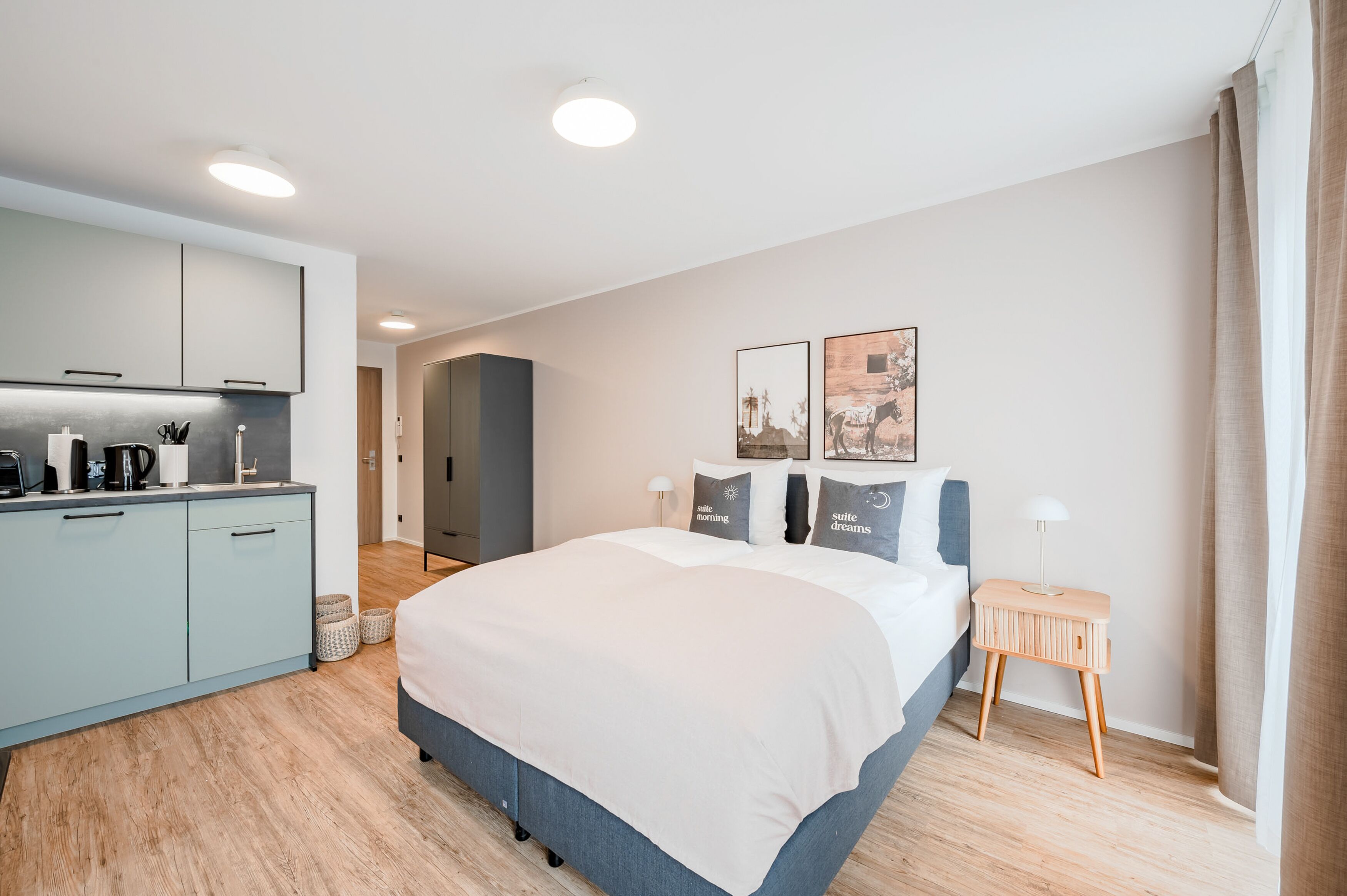 A modern hotel room with a large bed, a kitchenette, and a small side table.