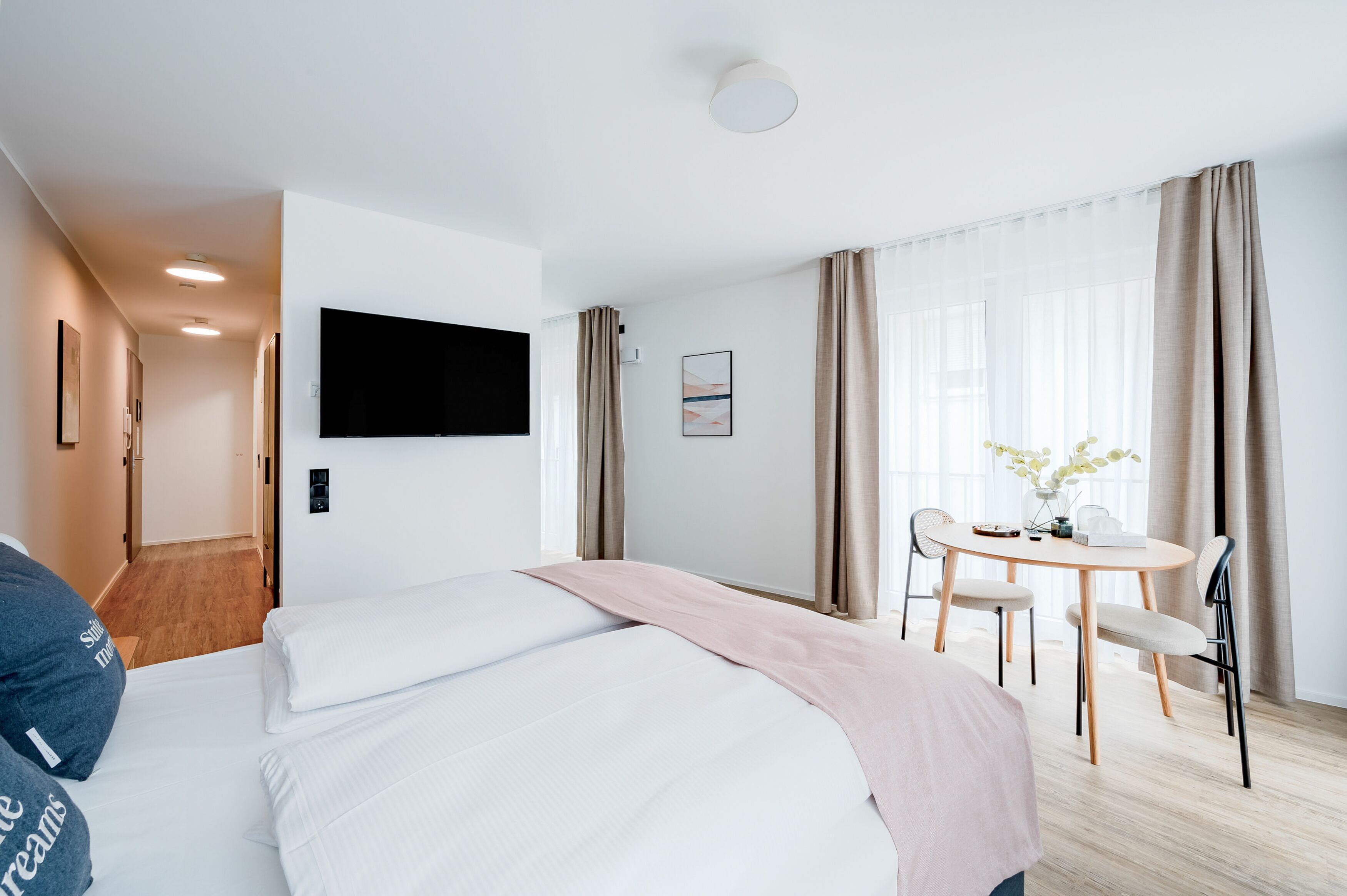 Une chambre d’hôtel avec un lit, un bureau et une chaise. Il y a un téléviseur fixé au mur et une fenêtre avec des rideaux.