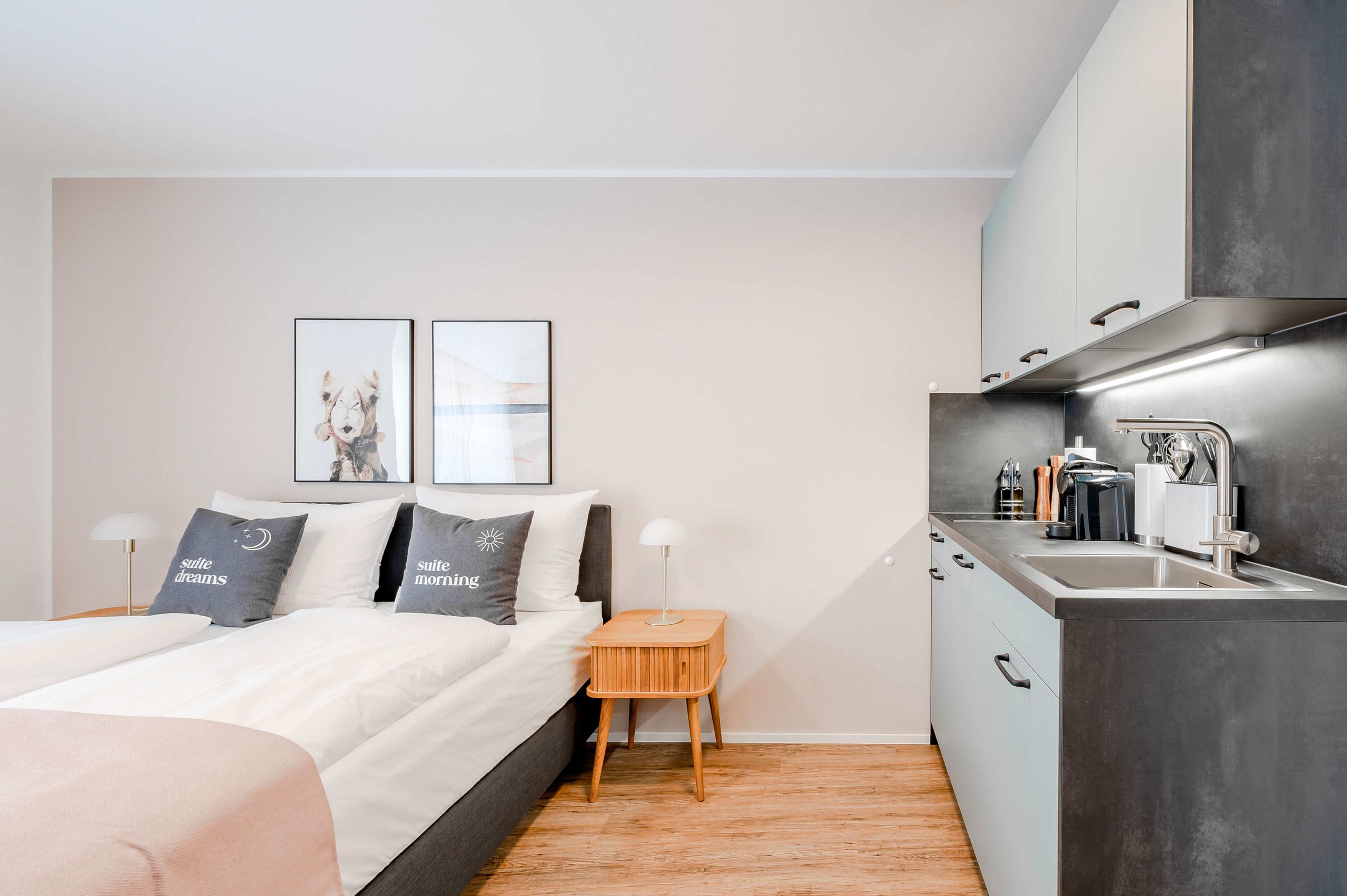 A modern bedroom with a bed, bedside tables, a small wooden stool, and a kitchenette with a sink and cabinets.