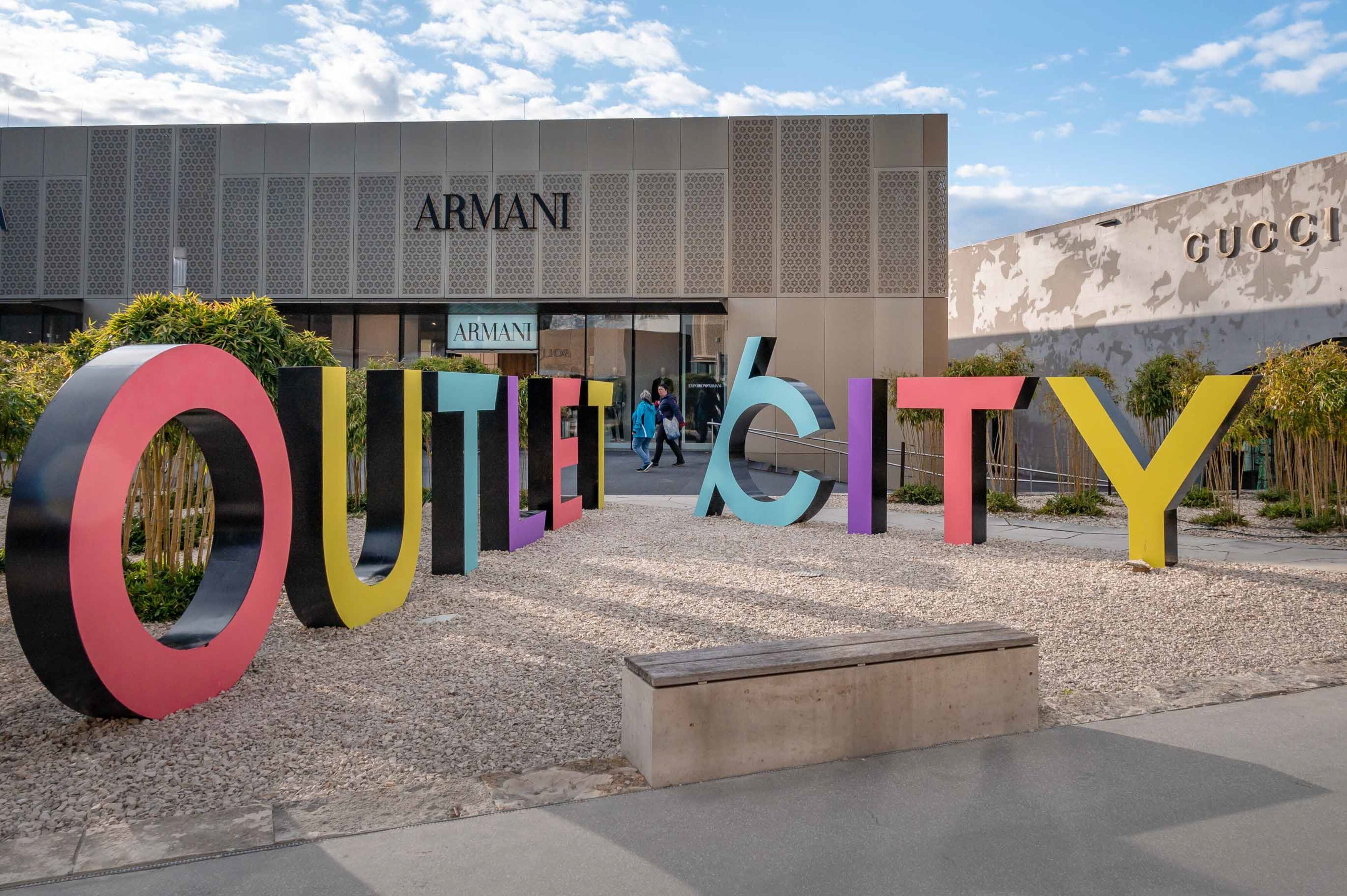 Ein modernes Gebäude mit den Hinweisschildern 'ARMANI' und 'GUCCI', umgeben von farbenfrohen, großen Buchstaben, die 'OUTLOOK' buchstabieren.