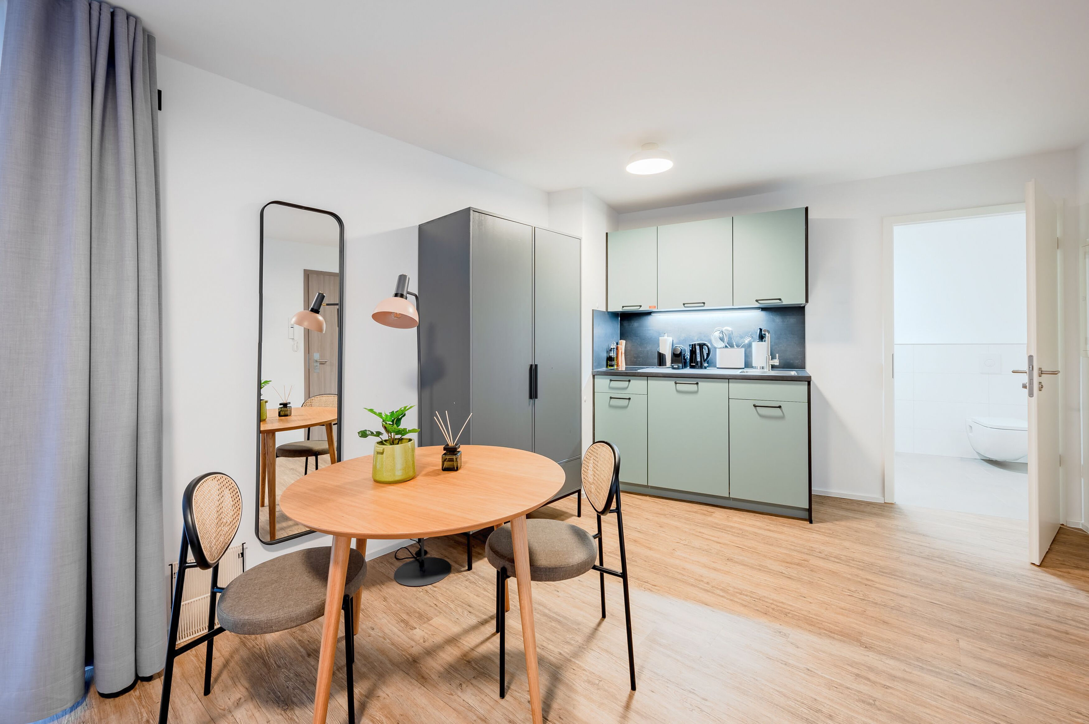 A modern kitchen with light green cabinets, a round wooden table with two chairs, and a large mirror above the table.