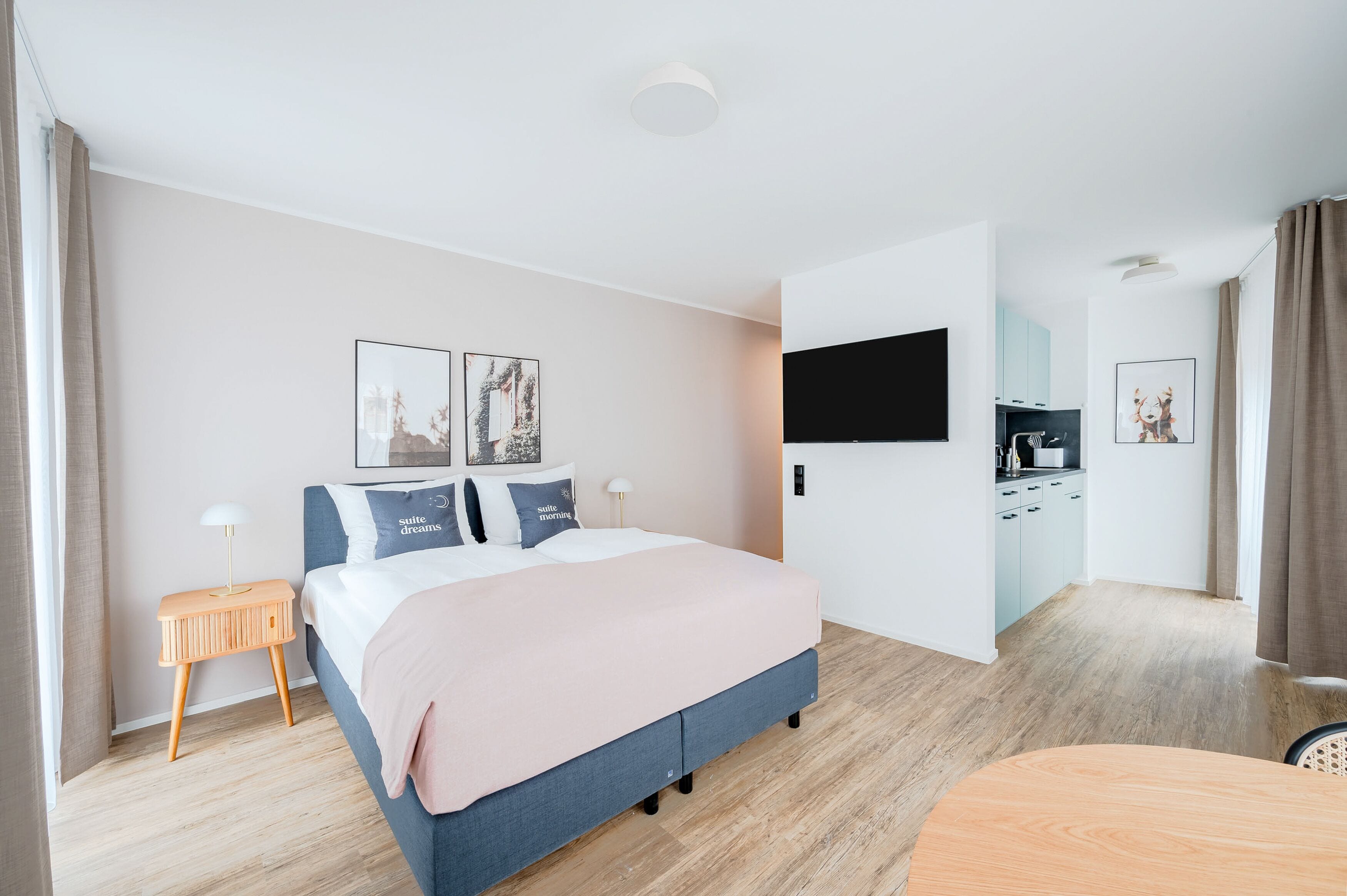 A modern hotel room with a bed, bedside table, TV, and a kitchenette area.