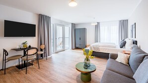 Una habitación de hotel moderna con sofá, una mesa de comedor con sillas, televisor de pantalla plana montado en la pared, cama con almohadas y un gran ventanal con cortinas.