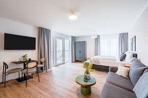 Quarto de hotel moderno com sofá, mesa de jantar com cadeiras, televisão de tela plana presa à parede, cama com travesseiros e uma janela grande com cortinas.