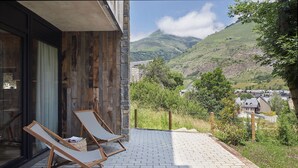 A balcony with two wooden lounge chairs, a stone wall, and a view of a village and mountains.