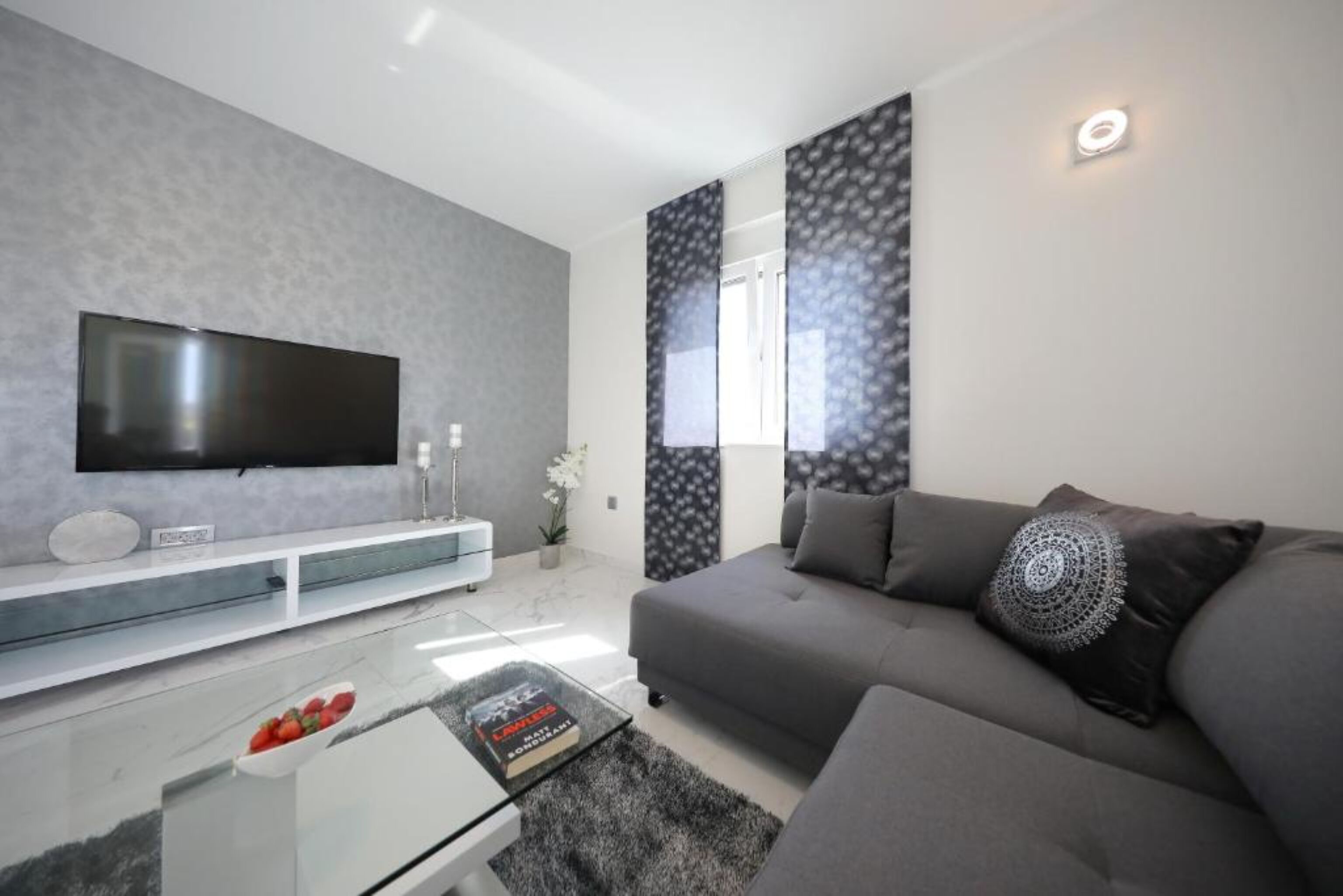 A modern living room with a flat-screen TV, grey sofa, and a glass coffee table with a book.