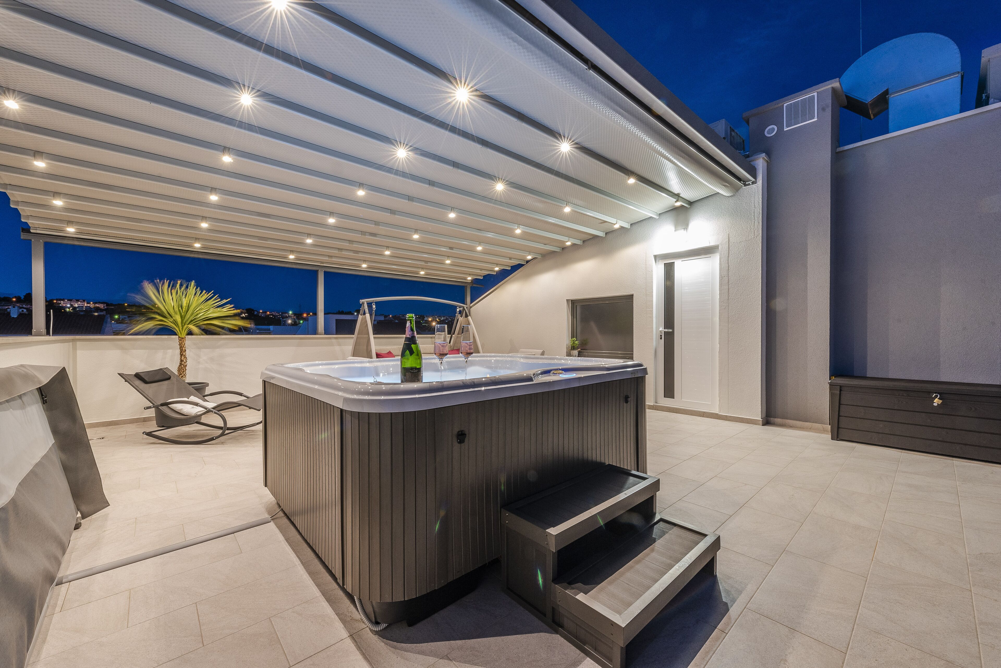 A modern outdoor hot tub area with a covered patio, lounge chairs, and a view of the city at dusk.