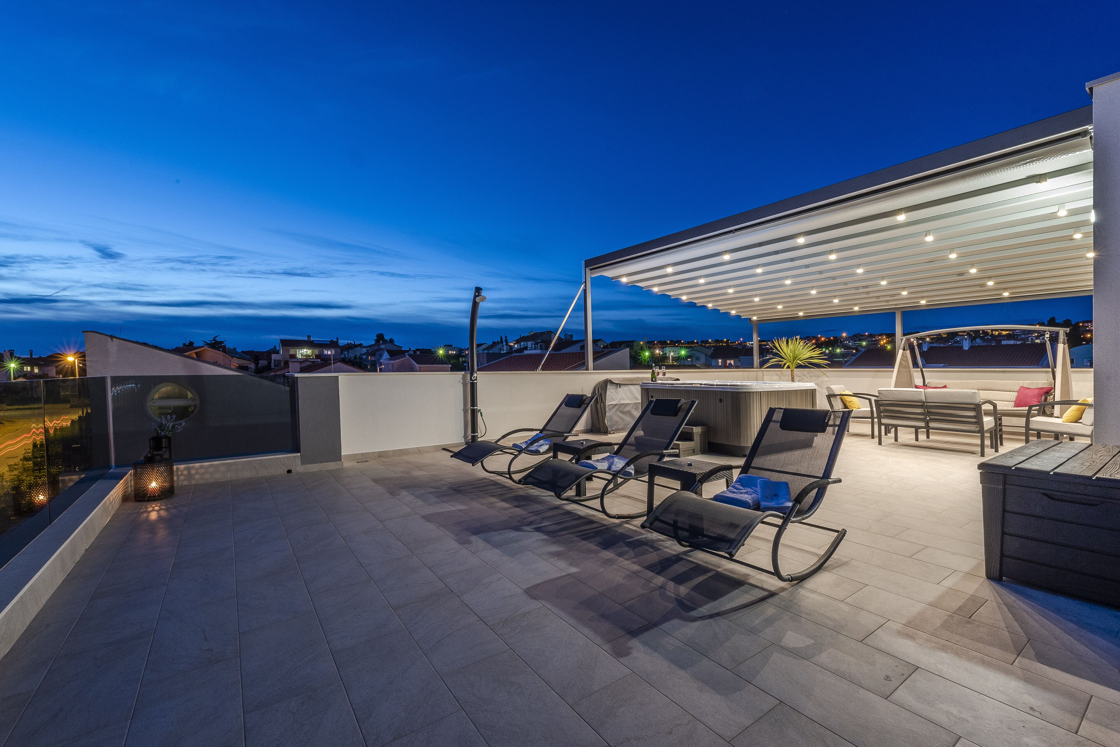 A rooftop terrace with a covered seating area, a grill station, and a view of the cityscape at dusk.