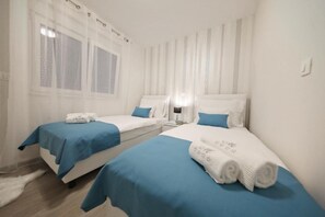 A hotel room with two beds, each with a blue bedspread and white pillows. There is a window with curtains, a nightstand with a lamp, and a white rug on the floor. - Luxury Majpruz Suites I (Zadar)