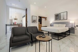 A modern hotel room with a bed, two armchairs, a small table, a desk, and a kitchenette.