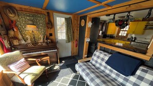 A cozy cabin interior with a piano, a checkered sofa, a dining area, and a kitchen. - Nature Retreat, Pet Friendly Cozy Cabin On 80 Acres With Fields & Forrest Trails (Fort Edward)