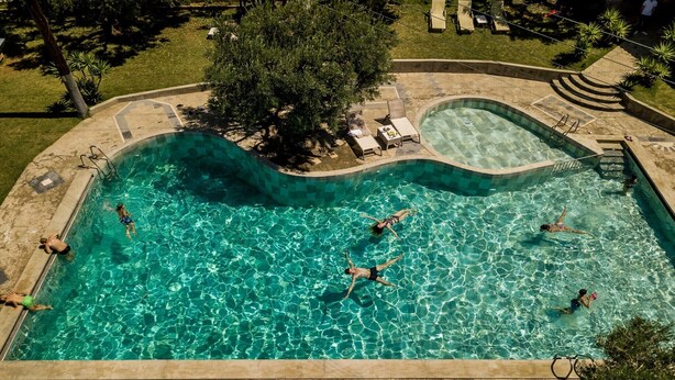 En pool med en böjd design, omgiven av en stenbelagd terrass och utomhusmöbler.