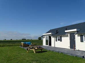 A white building with a dark roof, a patio with wooden furniture, and a grassy area. - Chalet for 4 persons in Kamperland, close to beach and sea (kamperland)