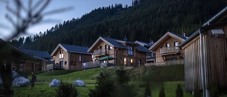 Filas de cabañas de madera con balcones, con un paisaje de ladera boscosa de fondo.
