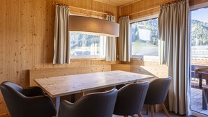 Interno di una baita in legno con tavolo e sedie, ampie finestre con tende e vista su una zona boschiva.