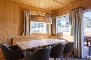 A wooden cabin interior with a dining table and chairs, large windows with curtains, and a view of a forested area.