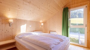 Camera da letto in una baita di legno, con un letto, due luci a parete, una finestra con tenda verde e vista su una foresta.