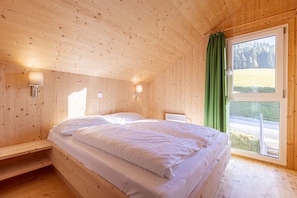 A wooden cabin bedroom with a bed, two wall-mounted lights, a window with a green curtain, and a view of a forest.