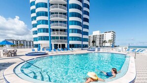 A swimming pool with two people, surrounded by lounge chairs and a building with balconies.