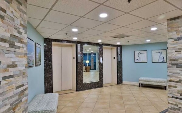 A hotel lobby with a tiled floor, stone accent walls, framed artwork, a bench, and a mirror reflecting the interior.