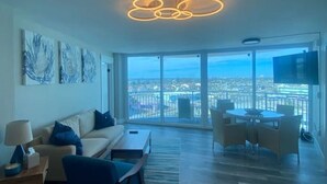 A modern living room with a sofa, coffee table, and dining area. There is a large window offering a city view.