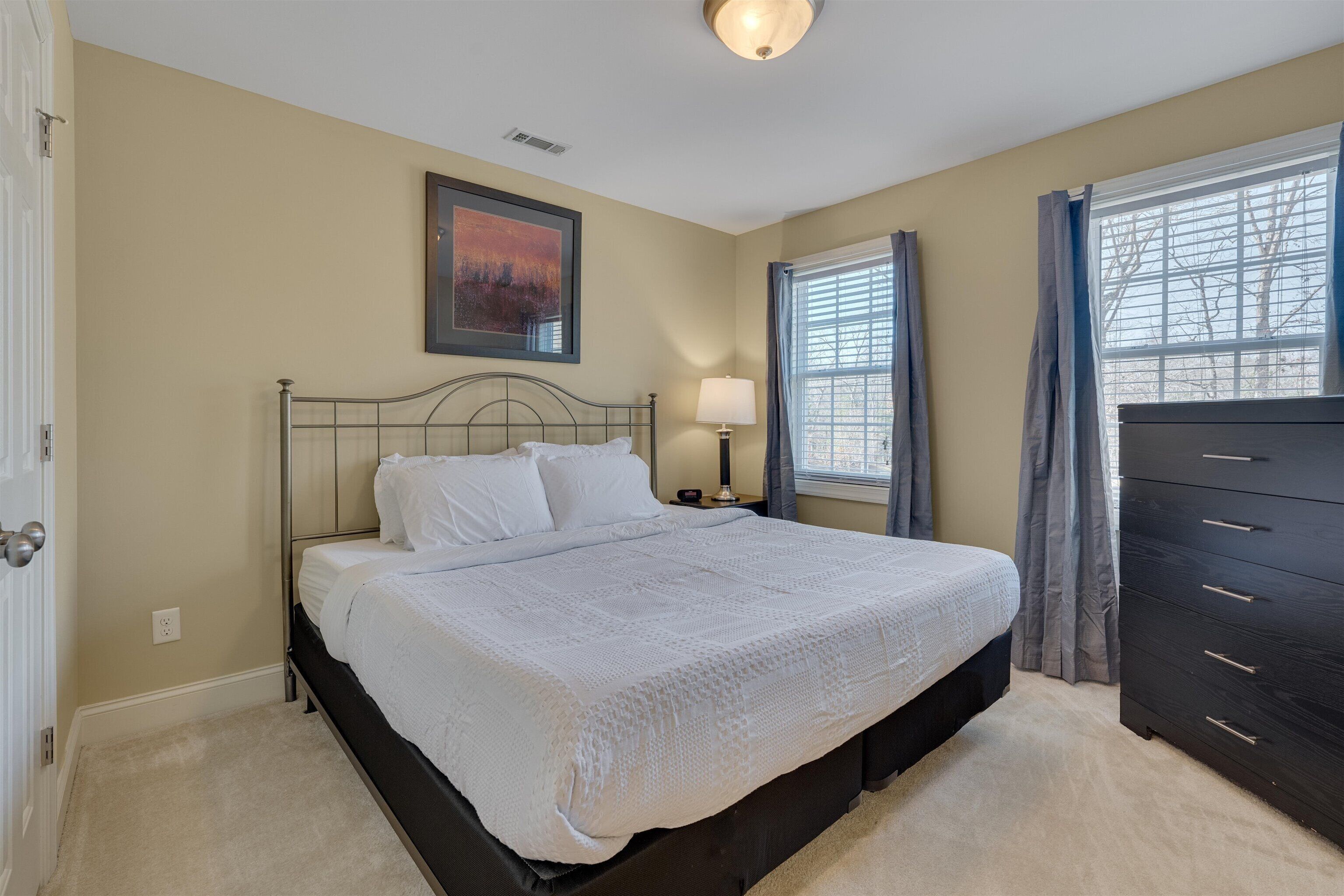 A bedroom with a large bed, a dresser, a nightstand with a lamp, and a window with curtains.