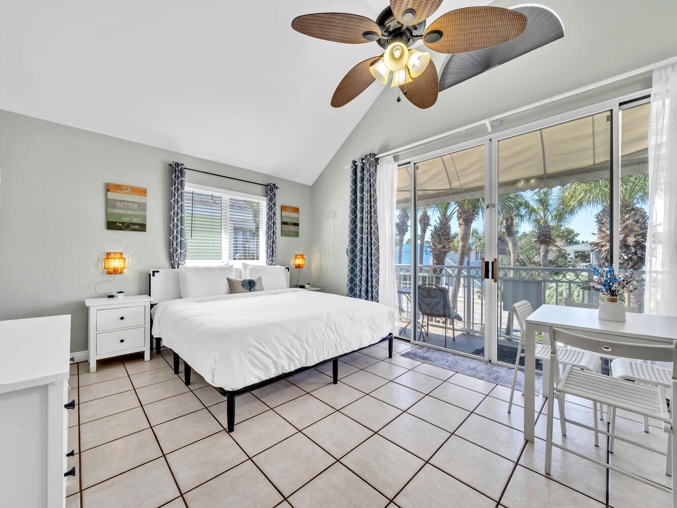 A bedroom with a bed, a nightstand, a desk, and a chair. There is a ceiling fan and a window with curtains.