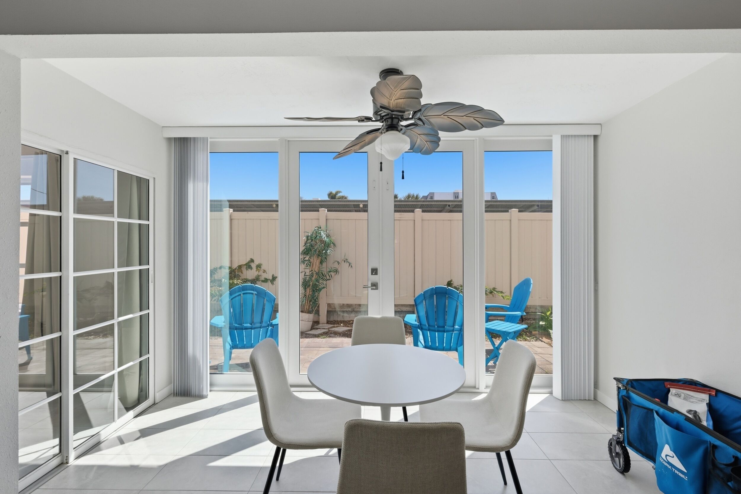 A room with a ceiling fan, a table with chairs, a sliding glass door leading to an outdoor area with blue chairs, and a blue luggage cart.