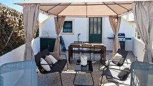 Zona d'estar a l'aire lliure coberta, amb taula, caires, una barbacoa i vista a un edifici amb persianes.