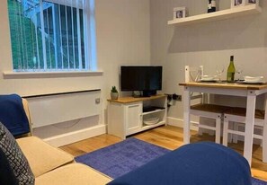 Un salon avec un téléviseur, une petite table sur laquelle se trouve une bouteille, et une fenêtre avec des stores.