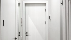 Un couloir étroit, avec une porte blanche au fond, un paillasson noir et un crochet fixé au mur.