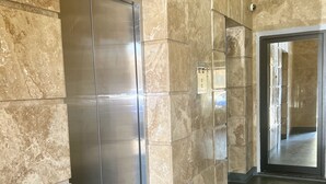 Un ascenseur moderne aux finitions métalliques, situé à côté d’un mur recouvert de grandes carreaux de marbre beige.