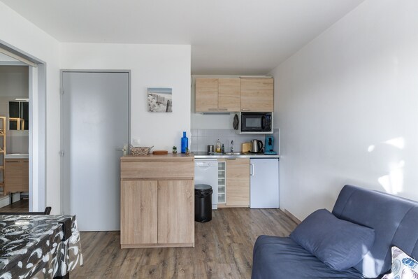 A compact kitchen with wooden cabinets, a microwave, and a dishwasher. There is a sofa and a table with a patterned tablecloth. - Océania -proche Mer et Thalasso (Cabourg)