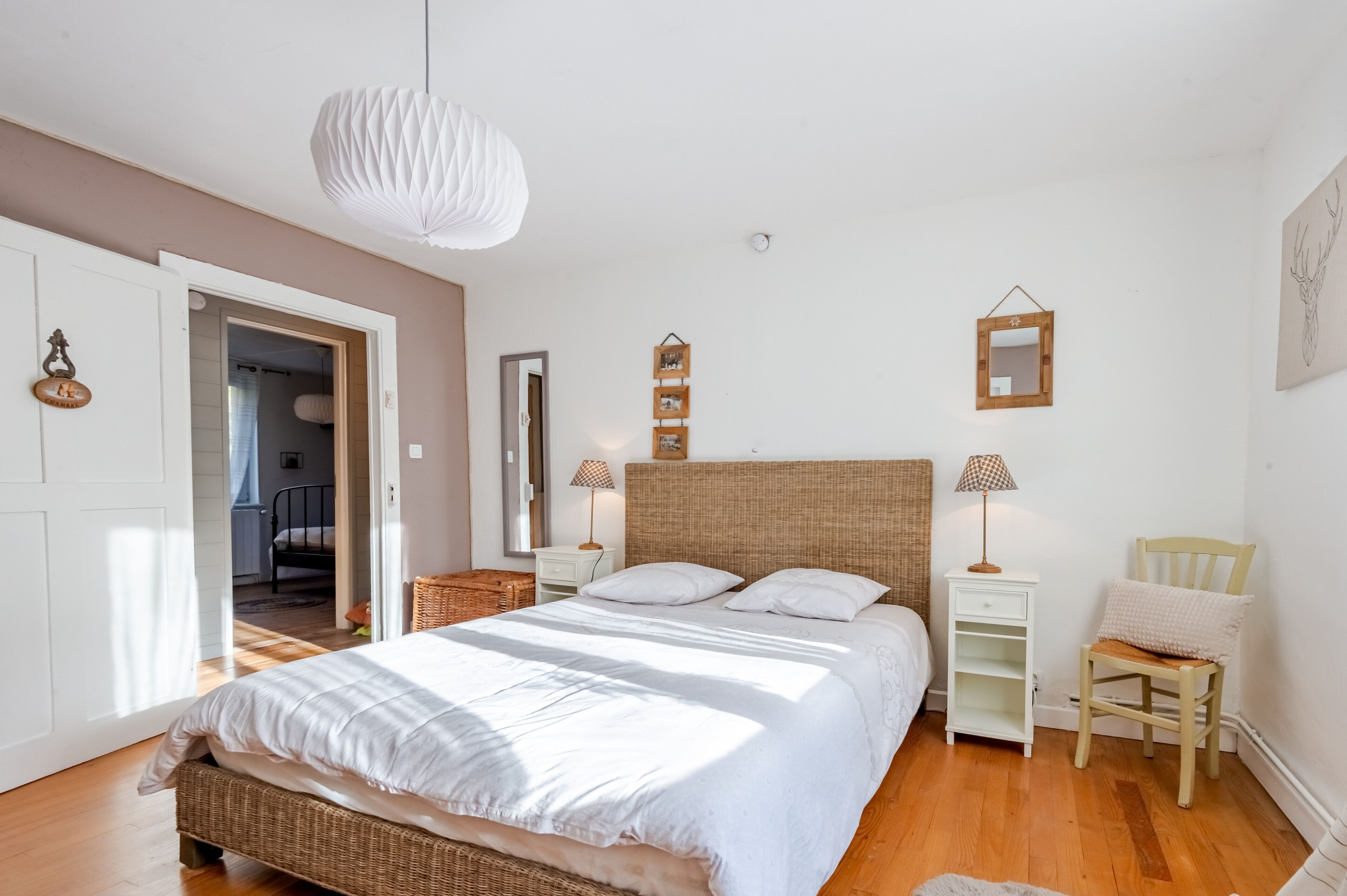 Un dormitorio con una cama grande, una mesita de noche, una silla, una lámpara colgante y una puerta que da a otra habitación.