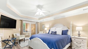 A bedroom with a white bed, blue bedding, a ceiling fan, a TV, a nightstand, and a chair.