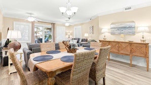 A dining area with a wooden table, wicker chairs, and a chandelier. There is a sofa and a cabinet in the background.