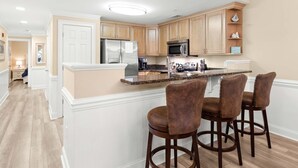 A modern kitchen with wooden cabinets, a stainless steel refrigerator, a microwave, and a granite countertop. There are three bar stools with cushioned seats.
