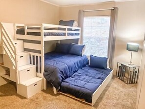 Una cama litera con un colchón y almohadas azules, una mesita con una lámpara y una ventana con cortinas.