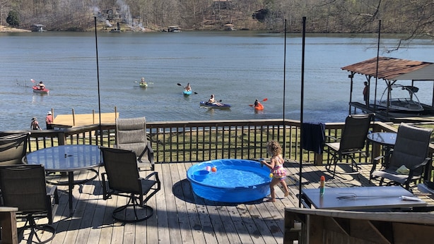 Une terrasse en bois avec coin salon extérieur, un jacuzzi et une vue sur des personnes pratiquant le kayak sur un lac.