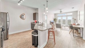 Una cocina moderna con encimera de granito, electrodomésticos de acero inoxidable y una pequeña isla con fregadero. Hay una zona de comedor con una mesa y sillas, y un amplio ventanal que proporciona luz natural.