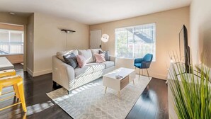 Een moderne woonkamer met een bank, salontafel en een blauwe stoel. Er is een groot raam met zonwering, een plant en een televisie.