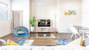 A modern living room with a flat-screen TV, a glass coffee table, a blue chair, a dining area with white chairs, and a potted plant. - Comfy 2BR king suite, fast WiFi, 8 min to Downtown (Austin)